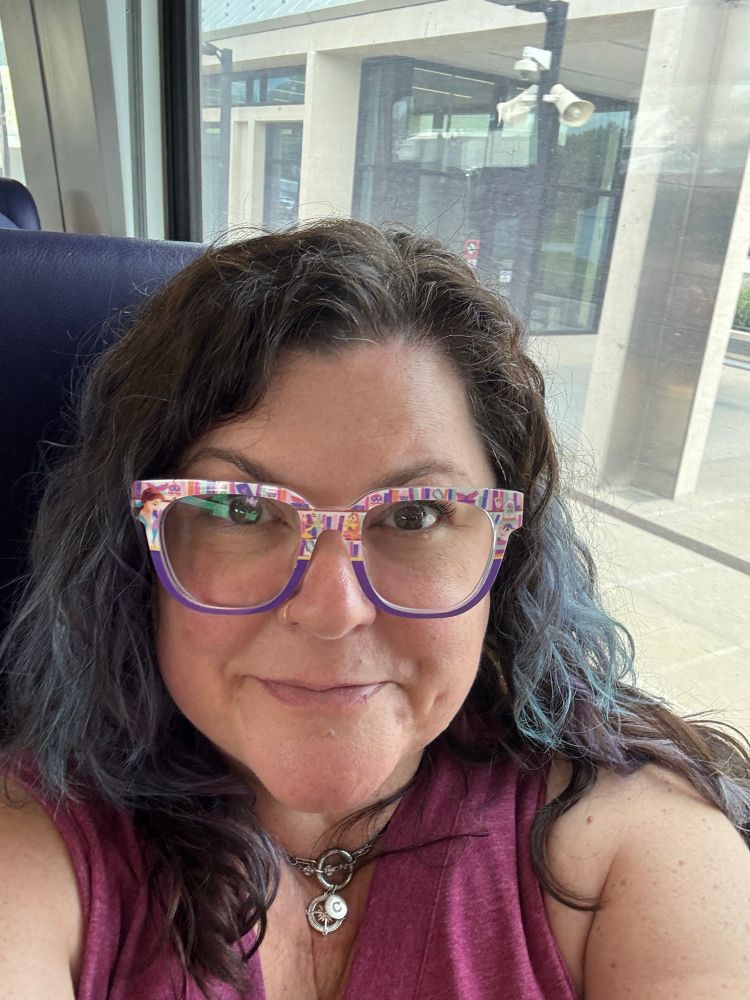 A middle-aged bespectacled brunette with some blue-ish, purple-ish hair smiles at the camera while riding Amtrak.