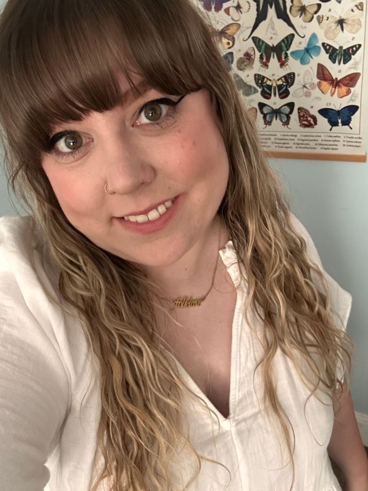 woman with brown and blonde hair wearing a white dress and “folklore” necklace, smiling in a selfie