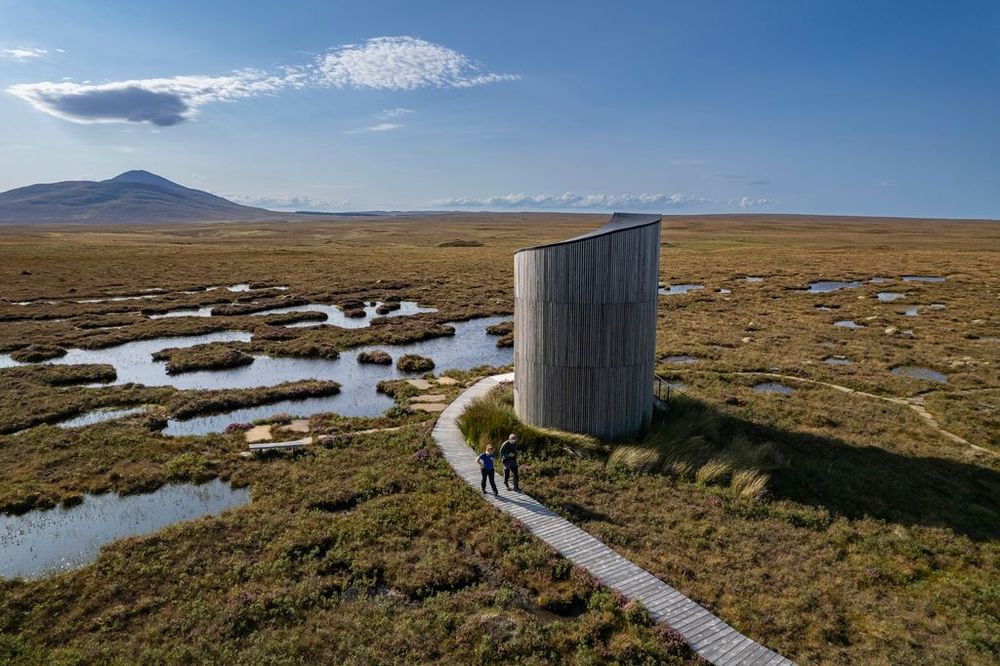 The cylindrical Flows Viewing Tower at Forsinard Flows Nature Reserve, Scotland, sits on a flat landscape of wet peatland which stretches to the hills on the horizon. Two people walk the Dubh Lochan Trail back from the tower.