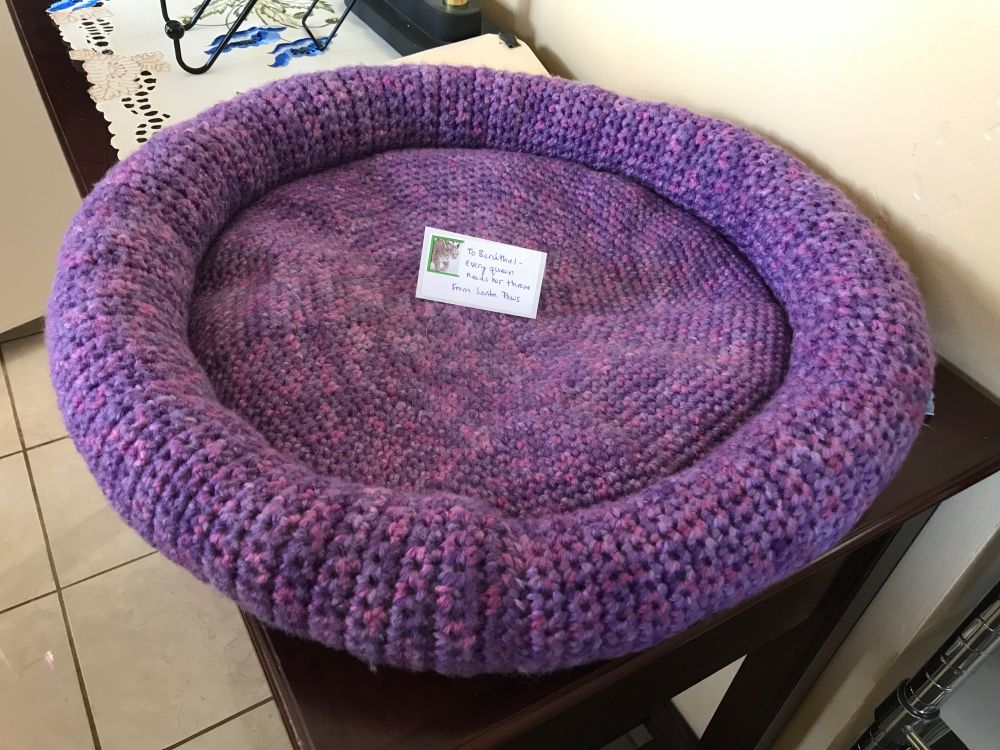 A circular crochet cat bed. The yarn is soft variegated purple and it's slightly lumpy so you know it's homemade. There's a gift tag in the middle reading "To Berúthiel - every queen needs her throne. From Santa Paws."