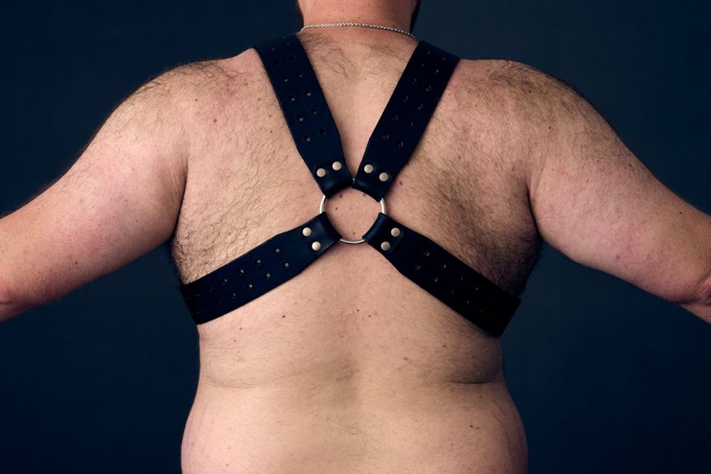Person with back turned to camera. They are wearing a leather harness with their arms spread to the side of their body