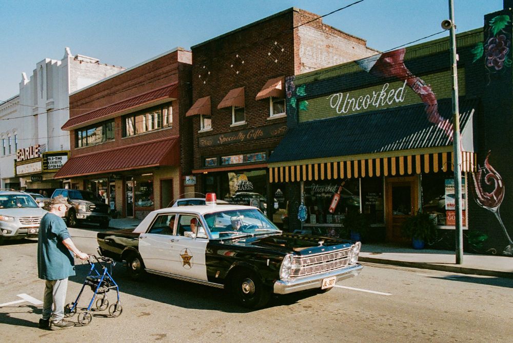 Old man waiting for an antique Ford police car to pass so he can cross the street.