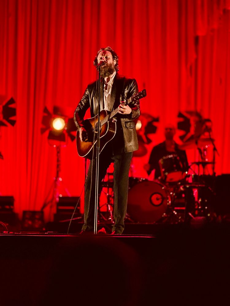 Father John Misty on a red background holding a guitar