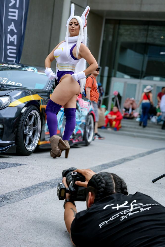 Another Mirko cosplay shot where I'm posing in a body building like pose standing with one bent up a little with both arms and hands flexed down. Behind me is an anime Itasca car and I'm posing for another photographer so this is almost like a behind thr scenes pic