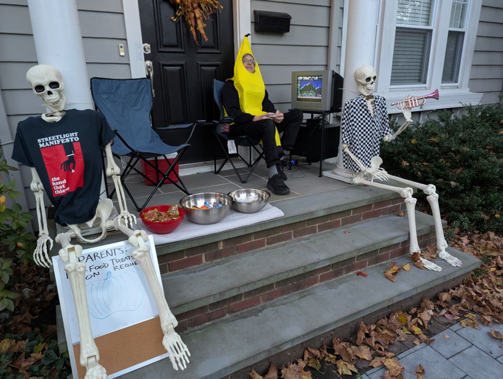 Matt wearing a banana costume, sitting on a folding chair on the front stoop. He's got a CRT set up and playing Castlevania III. On the steps are two plastic skeletons, one is wearing a Streetlight Manifesto shirt, the other a checkered shirt.