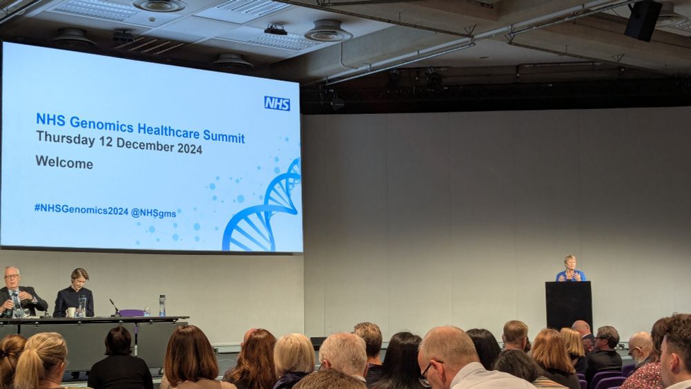 A packed conference hall, with Professor Dame Sue Hill introducing the Genomics Summit