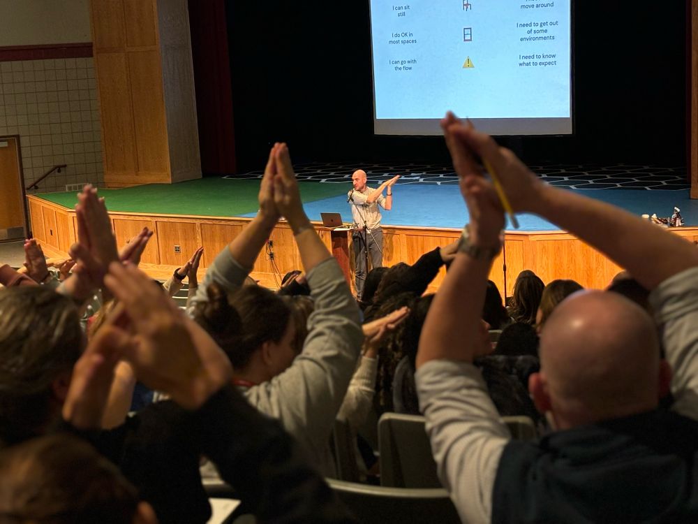 Aaron stands at a podium with his hands clasped over his head to the right and in the foreground dozens of workshop participants sit in their seats with their hands clasped above their heads, pointing in various directions
