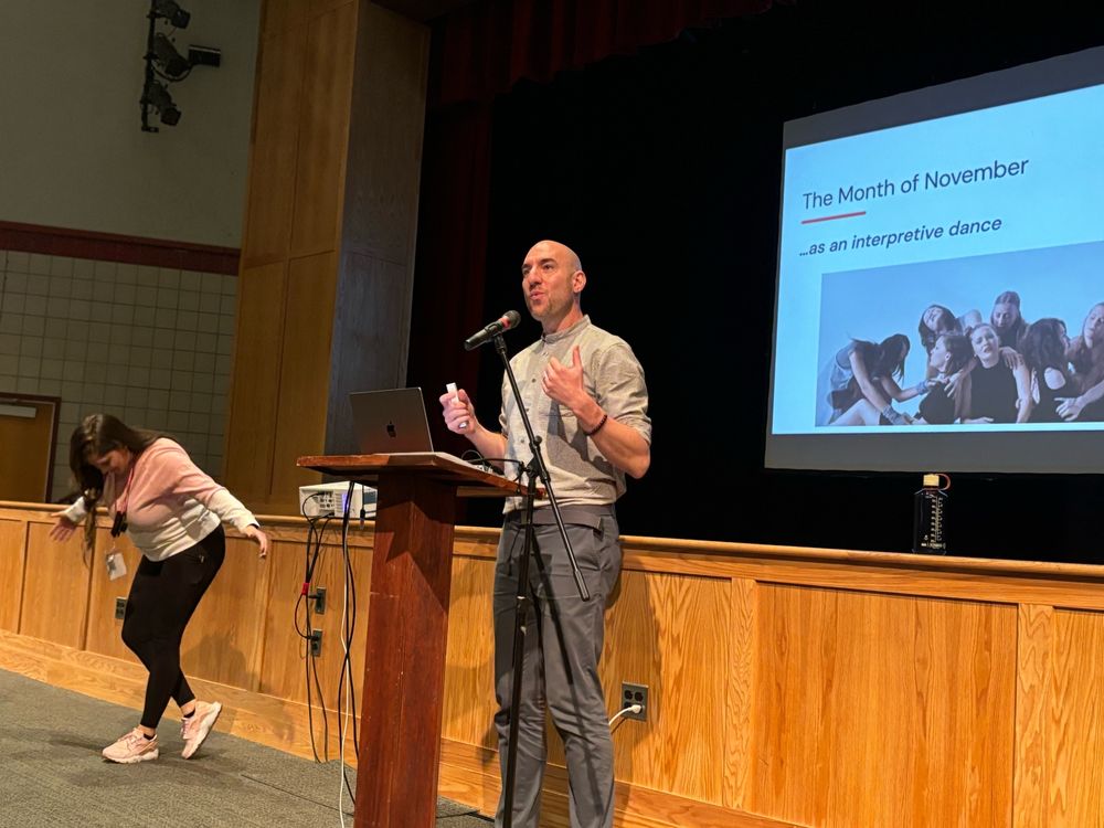 Aaron stands at a podium, talking into a microphone in front of a screen with a slide that reads an interpretive dance. next to him a workshop part participant takes a bow