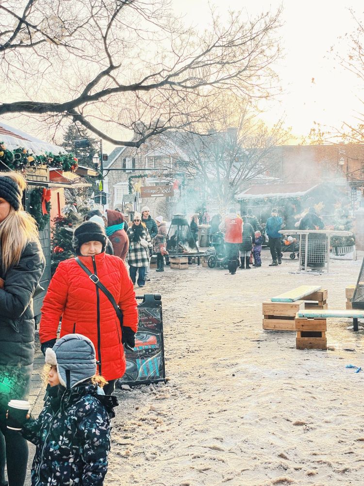An outdoor Christmas market covered in snow and smoke. People walk around to the various booths that are setup.