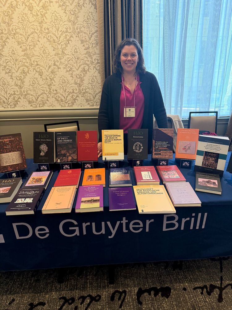 Woman standing behind book exhibit table with many books displayed. The background divided between beige wallpaper and a large curtained window. 