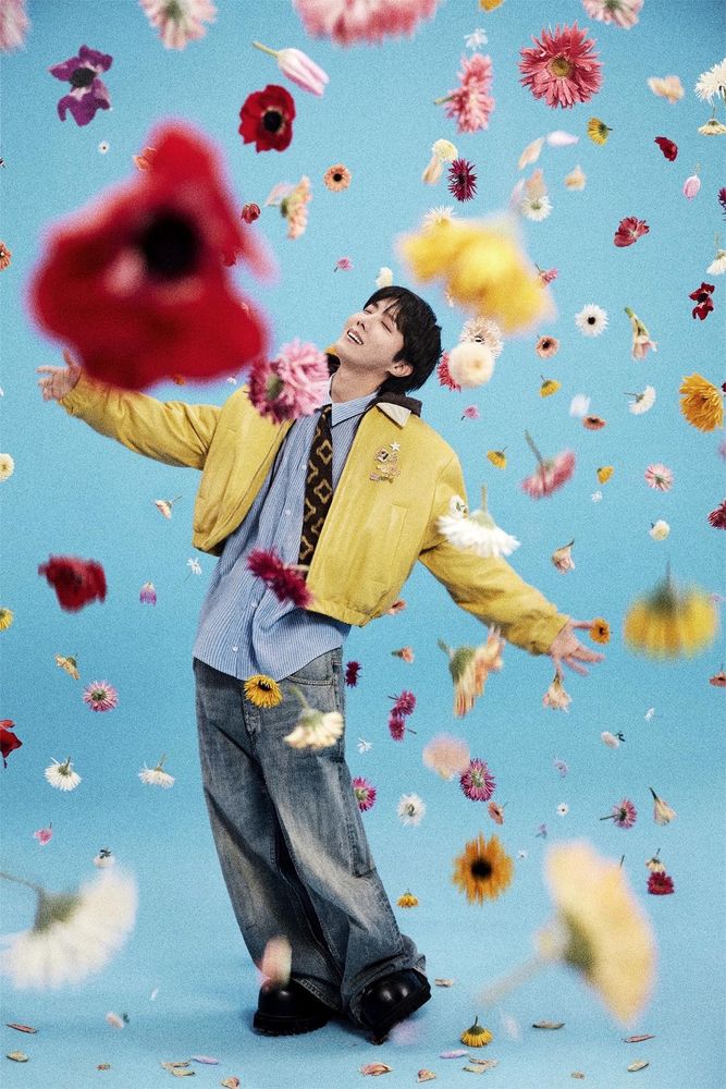 Hoseok with his arms spread and his eyes closed, wearing jeans, big black shoes, a blue button down, a brown patterned tie and a yellow jacket, standing in a rain of flowers.