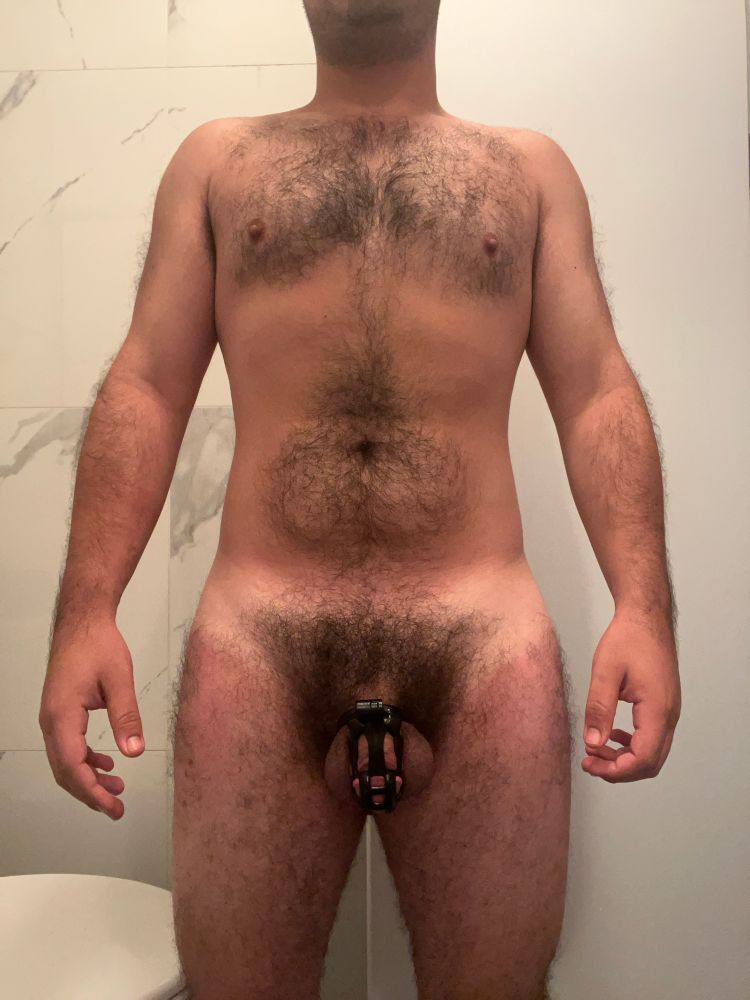 A furry man with a thick bush stands while wearing a black chastity cage