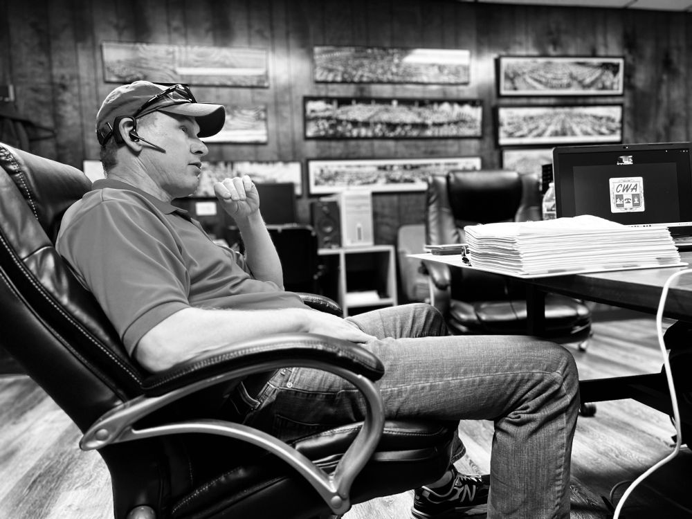 A man sits in an oversized chair at a desk in a wood paneled office. 