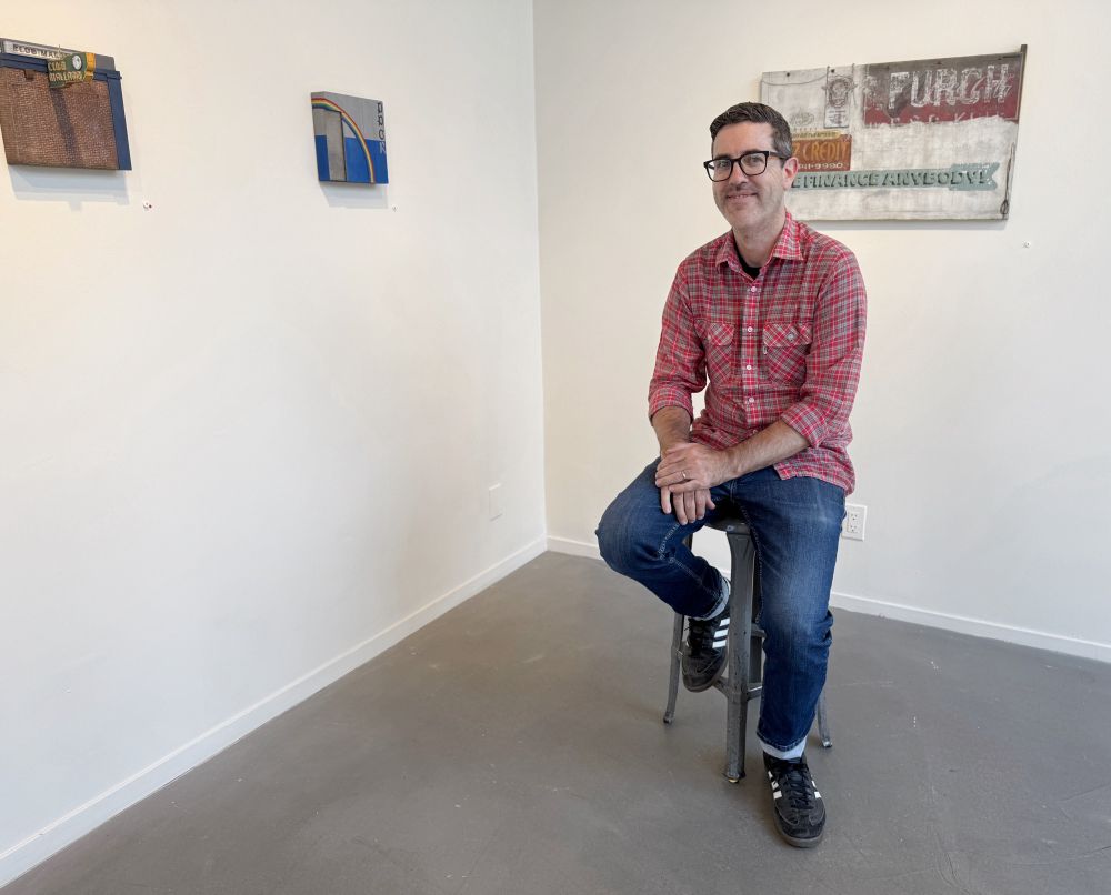 Artist Chris Rummell sitting on a stool in the gallery surrounded by his artwork on the walls.