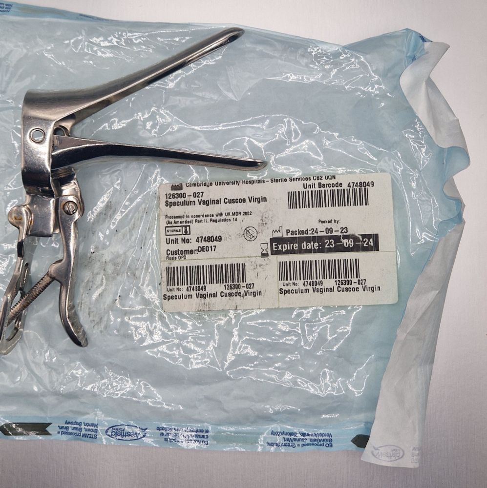 A metal vaginal speculum - an object with two blades open like the bill of a duck. It is resting on its packaging from Cambridge University Hospitals Sterile Services, labelled Speculum Vaginal Cuscoe Virgin. Packaged 24-09-23 Expires 23-09-24