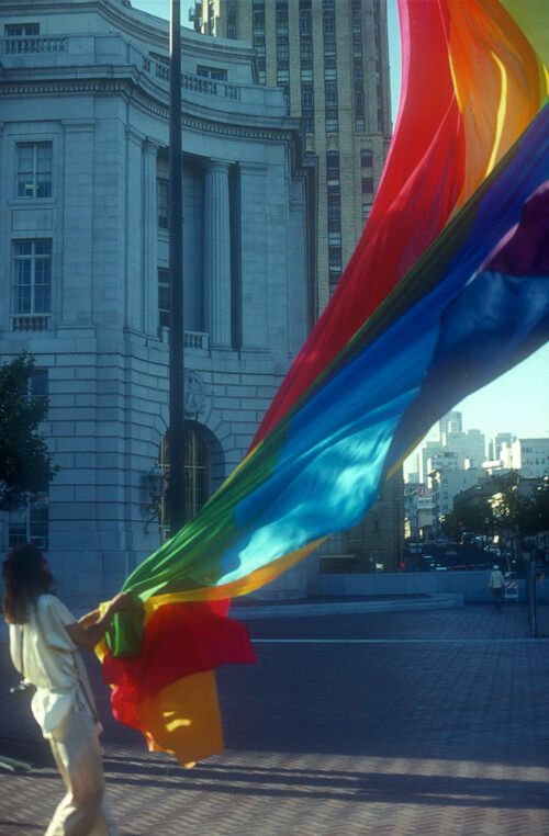 A person stands supporting the bottom of a huge flag being hoisted. The flag is a rainbow with eight stripes of pink, red, orange, yellow, green, turquoise, indigo and violet. 