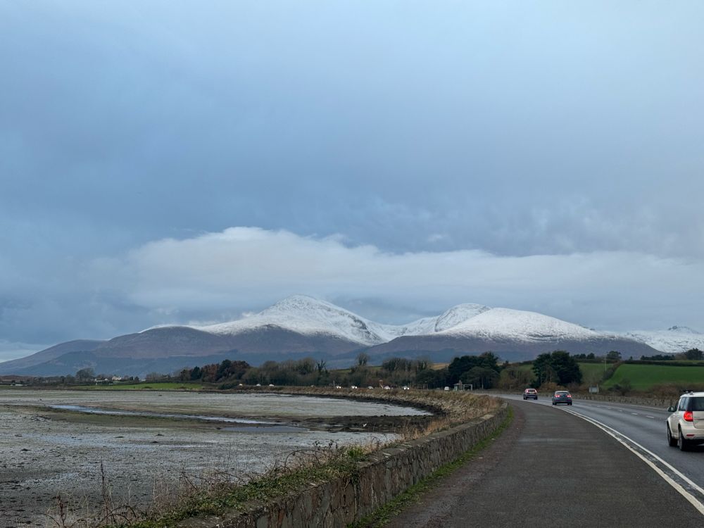 Image shows Mourne mountains with tops covered in snow. Clouds overhead and cars driving past