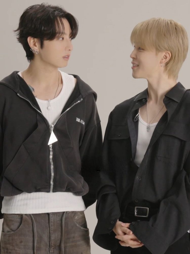 A screenshot from Jimin and Jungkook's GQ Japan q&a clip, right after Jungkook complimented Jimin for expressing himself well and Jimin asking if it was really okay. Jungkook is looking at Jimin, wearing a dark hoodie, white sleeveless top underneath. Jimin is looking at Jungkook with a beautiful smile, wearing a black button up not buttoned up, but tucked in his trousers, with a white shirt underneath, clasping his hands prettily in front 