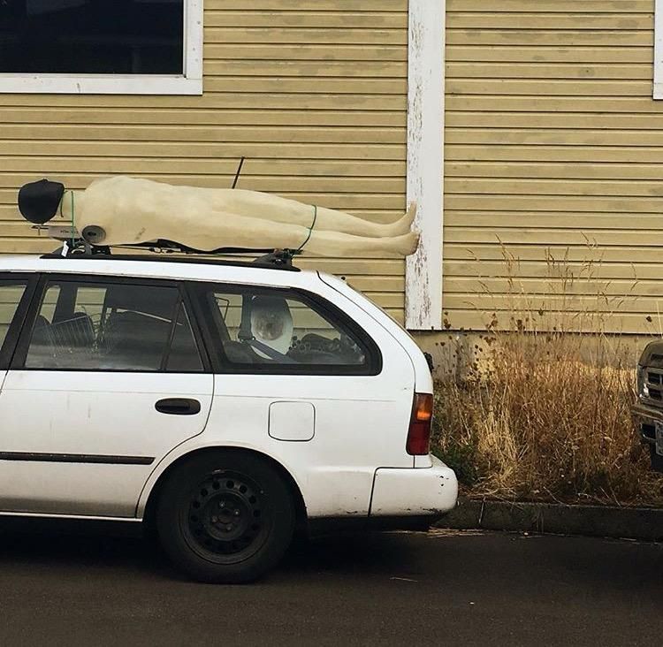 A plastic armless male mannequin strapped to the top of a white station wagon. 