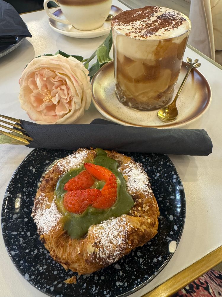 Matcha and strawberry crown (pastry with matcha cream and fresh strawberry) and a chestnut whisper latte served in a glass, with gold cutlery rolled in a black napkin. A large silk rose lies next to the saucer cradling the coffee glass. Both from the wonderful Tokyo Bagel. 