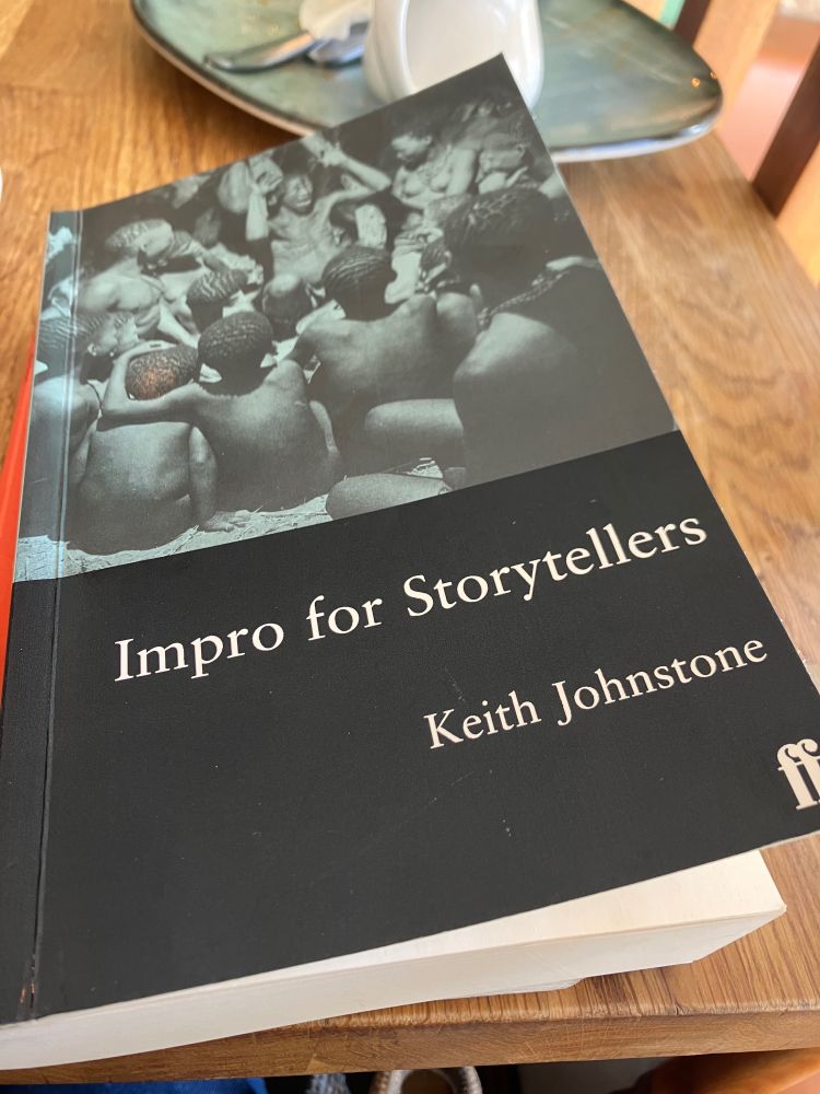 Close up of Impro for Storytellers by Keith Johnstone. Grey cover, writing at bottom, image above of a tribe of (African?) people watching someone who looks as if they are telling a great story