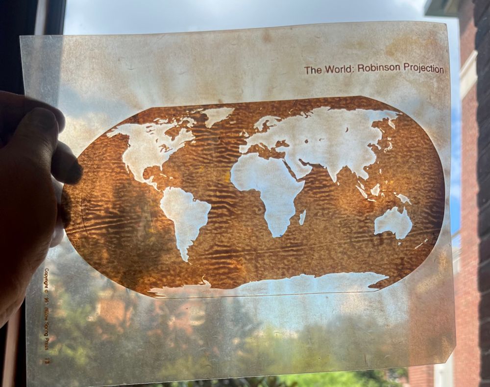 The photo shows a hand holding an overhead projector transparency up to a window. The image on the transparency is a map of the world.