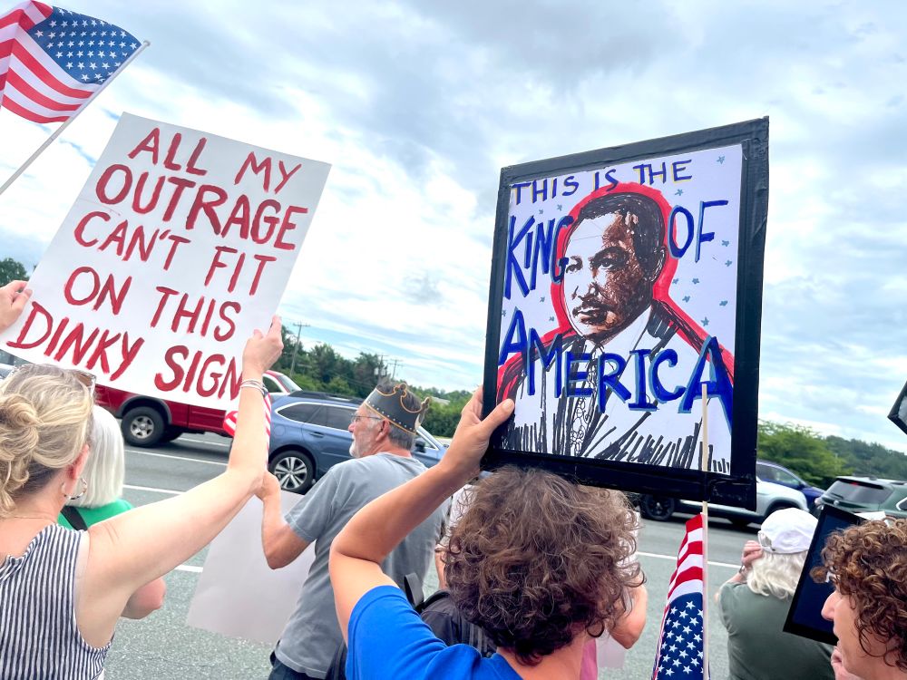 The photo shows two white women, who are members of a large crowd of protesters. Although they are seen from behind, their signs can be read. One shows a photo of Martin Luther King and says, "This is the King of America." The other sign reads, "All my outrage can't fit on this dinky sign."