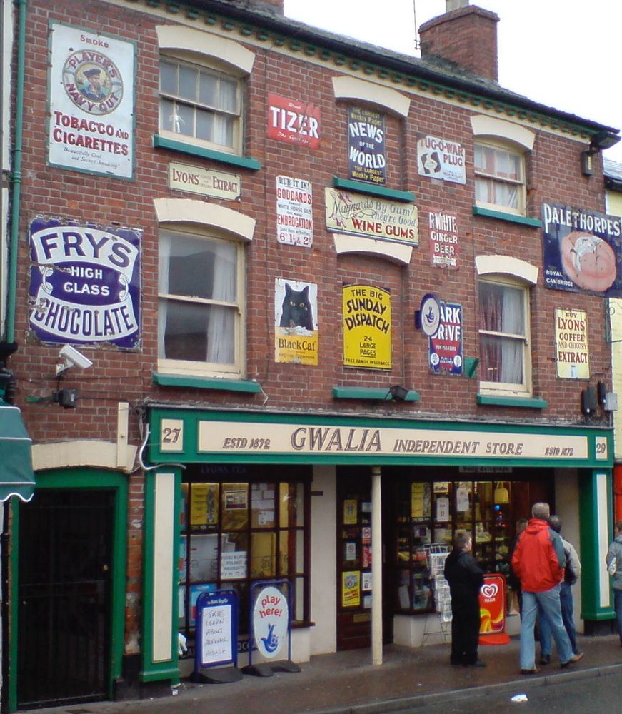 Shopfront with old signs above advertising brands such as Fry’s chocolate, Tizer, Palethorpe’s sausages, the Sunday Dispatch, R White’s ginger beer, etc. 