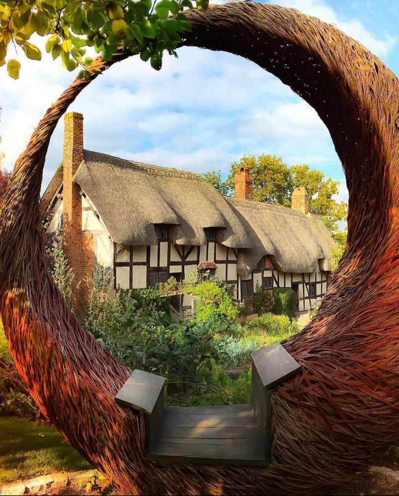 View through a beautiful curved wood structure to a long thatched cottage with three chimneys on top. Above the frame hangs an apple tree, ripe for the picking!