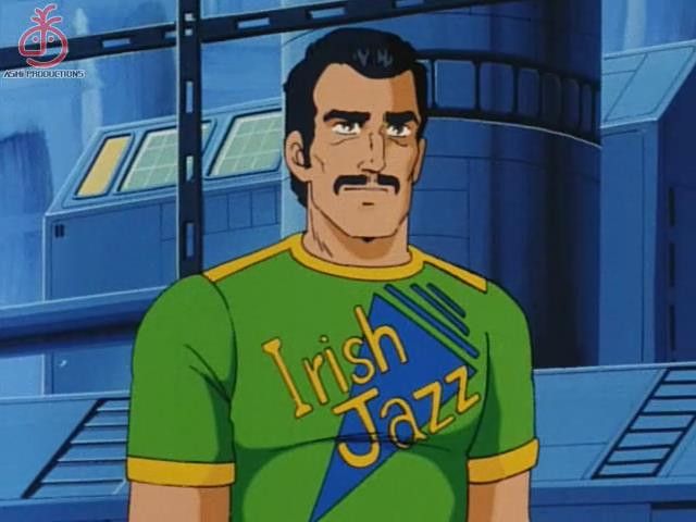 1980s anime screenshot of a square jawed man with olive skin, black hair and a large mustache in a futuristic metal backdrop with screens and panels in the background. He is wearing a green tshirt shirt with yellow piping at the collar and sleeve cuffs that has IRISH JAZZ on the front over a blue triagle with some lines on one side to made it 80s style 