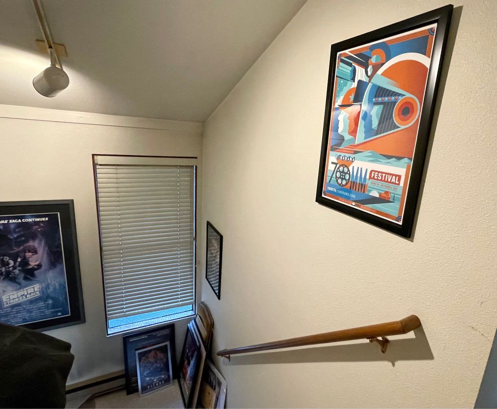 This is a photograph of a stairwell. The walls are cream colored. On the wall at the top of the staircase hangs a brightly colored poster in a black frame from the Cinerama‘s 70 mm Film Festival in Seattle. At the bottom of the staircase, a poster from the 1999 movie The Mummy hangs on the wall. There is an empty stretch of wall between them. Beneath the poster of The Mummy, several framed posters lean against the wall, yet to be hung.
