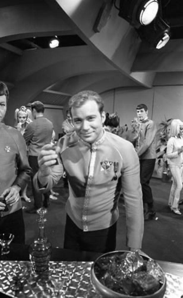 A black-and-white photograph of William Shatner as Captain Kirk in formal Starfleet dress uniform attending a party, people all around. He toasts the viewer.