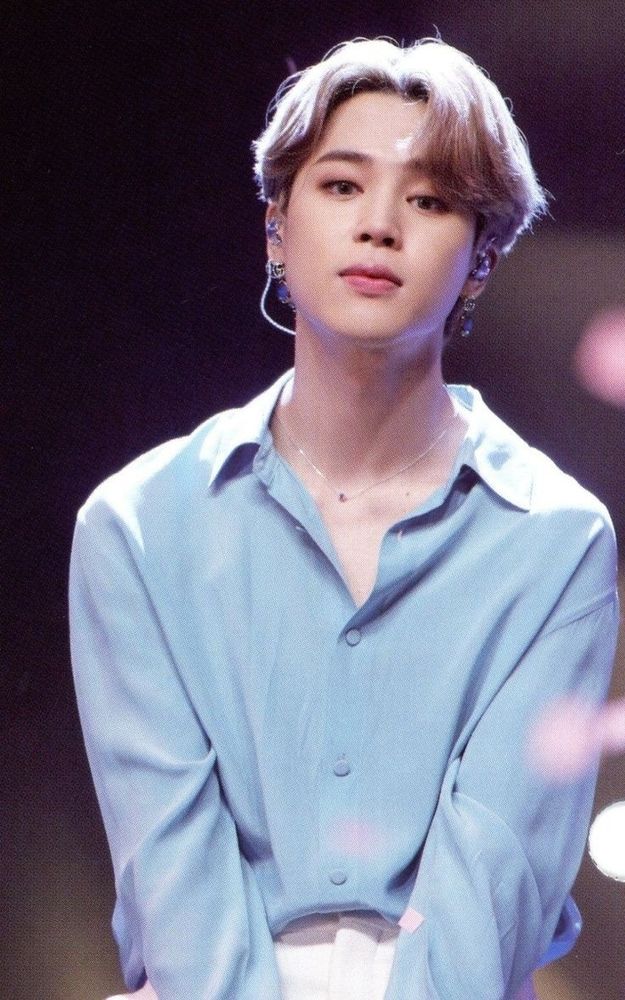 Blond Jimin sitting on a stool, wearing white trousers, a baby blue button up shirt, a silver necklace and silver earrings. His hair is styled in a middle part and he's looking directly at the camera. 