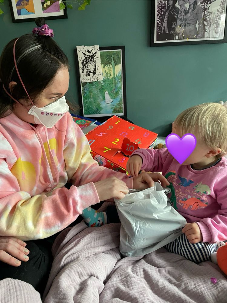 Fran a white woman with brown hair and a tie due hoody on, opens a bag with the 2 year old nephew who has blonde hair and a pink jumper 