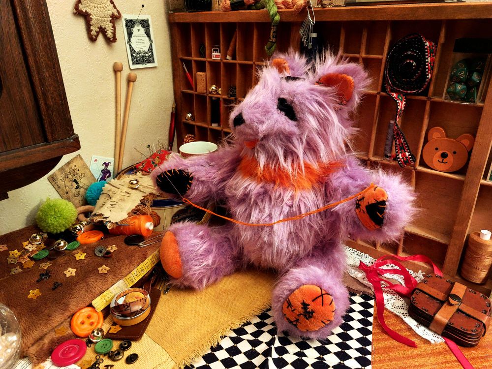 The Seam plush, still sitting down, but is now holding a needle threaded with orange embroidery floss in their paws.