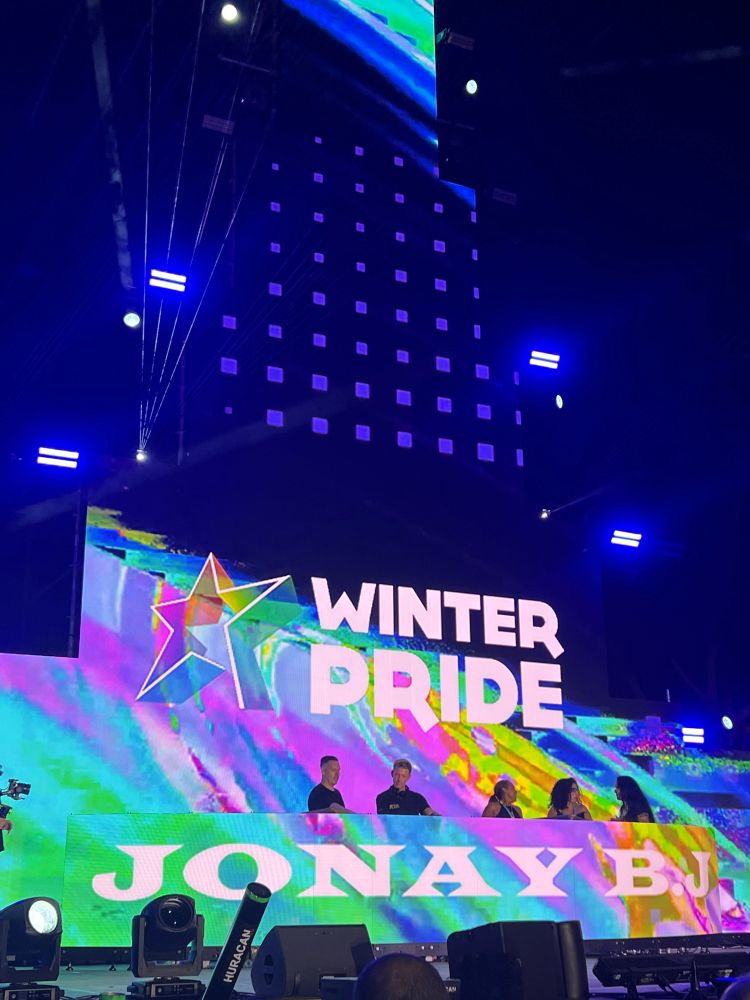 Die KI sagt zu diesem Foto:

📸 Bildbeschreibung:

• Bühne im Rampenlicht: Im Zentrum steht eine imposante Bühne mit riesigen LED-Screens, die in leuchtenden Farben das Logo WINTER PRIDE zeigen – flankiert von einem strahlenden, mehrfarbigen Stern, Symbol für Vielfalt und Zusammenhalt.
• Künstler*innen & Crew: Auf der Bühne sind mehrere Personen zu sehen – vermutlich Performer*innen, DJ oder Event-Team – eingebettet in dynamisches Lichtspiel aus blauen Spotlights und Laserstrahlen, die die Nacht in ein Kaleidoskop verwandeln.
• Musikalischer Moment: Der Name JONAY B.J erscheint auf einem der Screens – vermutlich der DJ oder Live-Act, der gerade die Menge zum Beben bringt.
• Atmosphäre: Die Szene wirkt wie ein pulsierender Herzschlag der Pride-Community – laut, stolz und voller Lebensfreude. Man spürt förmlich die Beats, die durch Maspalomas tanzen.
