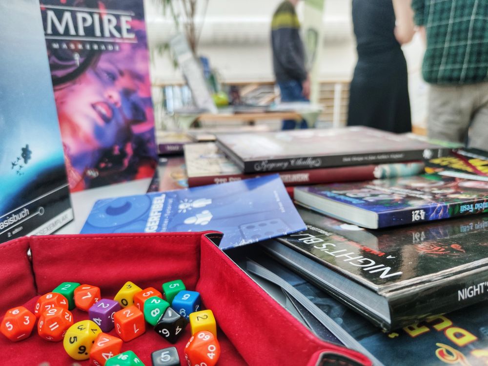 Detailbild eines Messestandes: Im Vordergrund verschiedene bunte Rollenspiel-Würfel, im Hintergrund sind unscharf Regelwerke verschiedenster Pen & Paper-Rollenspiele zu sehen.