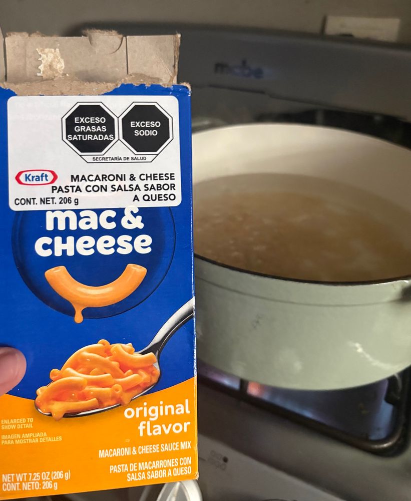 A box of Kraft Mac and Cheese (with excessive saturated fats and sodium level warnings) next to a sage green Dutch oven on a gas stove 
