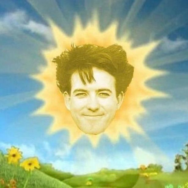 A humorous image edit featuring the bright yellow sun from the children’s show Teletubbies, with The Cure’s Robert Smith’s smiling face replacing the sun baby’s. The background shows a blue sky, soft clouds, and green hills dotted with yellow flowers.