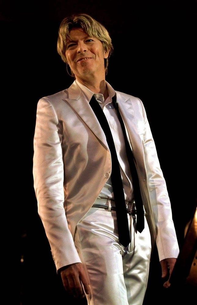 David Bowie smiling warmly on stage, wearing a shiny white suit with a white shirt and a loosely knotted black tie. His blond hair is styled with soft layers that frame his face. The lighting highlights the sheen of the suit against a dark background. 
I’m not sure who took this photo?