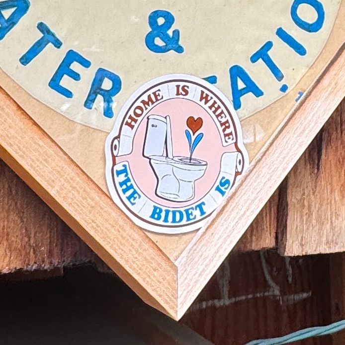 Close up of the “home is where the bidet is” sticker tucked into a sign frame. 