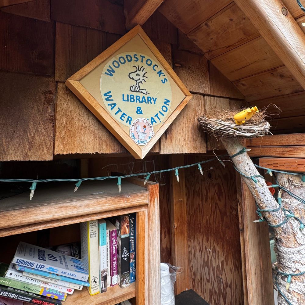 Sign that says “woodstocks library and water station”. A “home is where the bidet is” sticker sits tucked into the frame of the sign. 