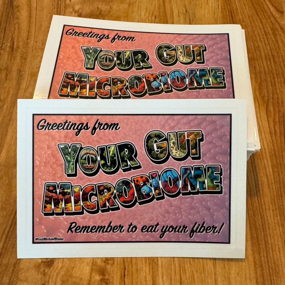 Photo of a stack of stickers made by Sticker Ninja (Portland, Or). The sticker is rectangular shaped with a white border. The sticker features text that says “greetings from your microbiome. Remember to eat your fiber!” The ‘your gut microbiome’ text is formatted in the style of a vintage postcard with different gastrointestinal elements filled into the text. The background is a pink and purple view of inside a gastrointestinal tract showing villi. 