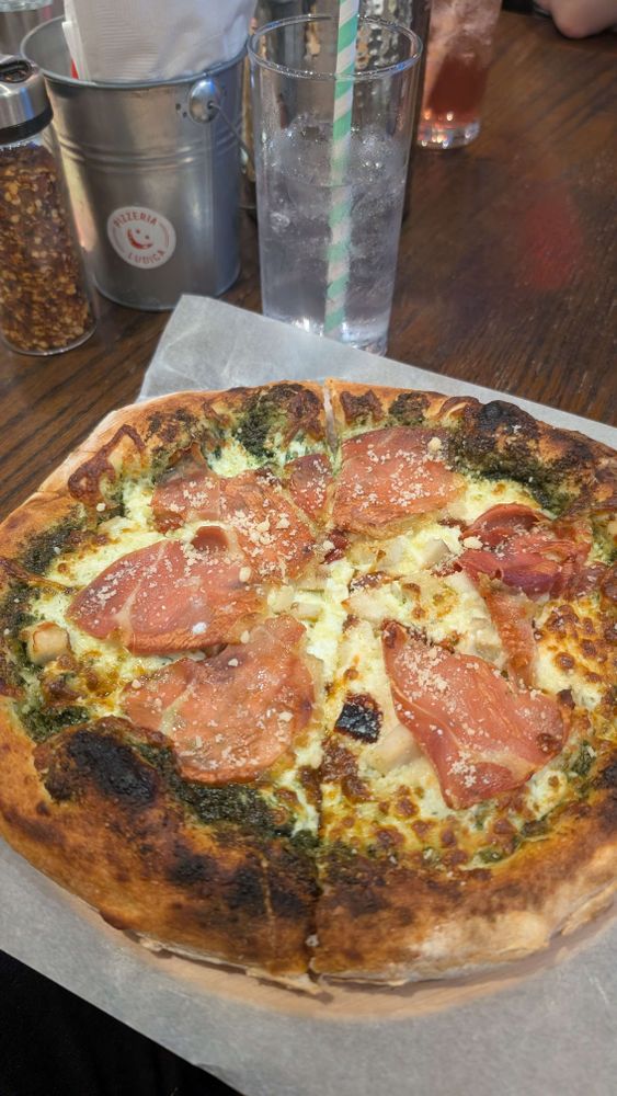 A woodfired chicken prosciutto and pesto pizza with a lavender soda in the background