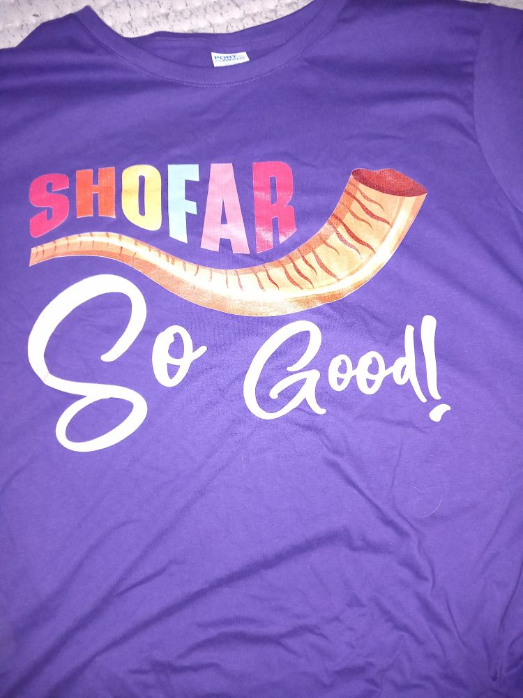Auf einem lila T-Shirt steht über einem in orangen und braunen Farben gezeichneten Widderhorn der Schriftzug "Shofar" in regenbogenfarbigen Buchstaben, darunter steht in weißen Buchstaben "So good".
Es ist eine Anspielung auf "So far, so good".
Das Shofar wird an hohen jüdischen Feiertagen geblasen und im Monat Elul, der der letzte Monat im jüdischen Kalender ist.
Rosh ha-Shana ist das jüdische Neujahrsfest.