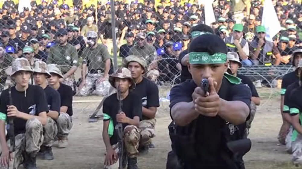 Kinder azs Gaza in militärischer Kleidung und Waffen. Ein Kind mit grünem Stirnband auf dem etwas in arabischer Schrift steht, steht im Vordergrund und zielt mit einer Pistole direkt auf den Betrachter.