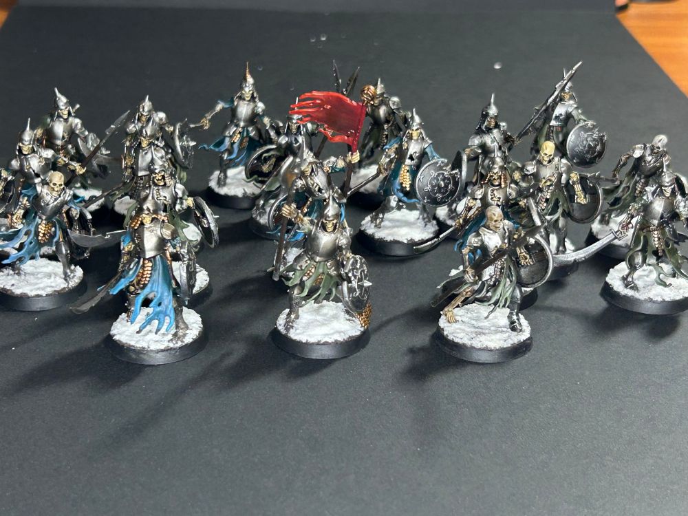 Collection of 20 skeleton warrior miniatures, painted in metallic silver and brass armors. They hold an assortment of swords and spears as they are sculpted as shambling forward, their bases with texture paint to look like a snowy tundra. Tattered green and blue cloth hangs from their forms and they hold silver shields painted to look tarnished.