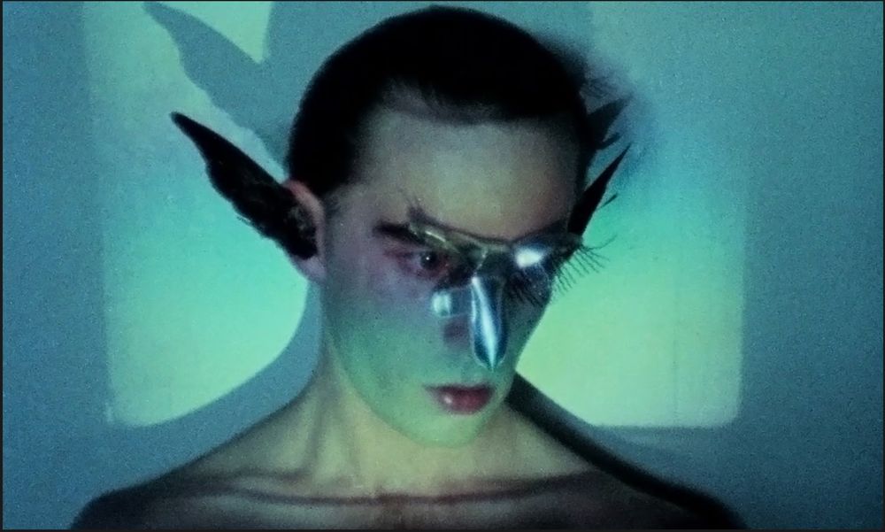 Image of a gender fluid artist Troy Vanessa Davies from the music video of the Hunters and Collectors song 'Talking to a Stranger' (description: a person in a bird-like mask with wings attached to their ears, shown from the shoulders up. The light on his/her face creates a rainbow pattern and a blue light shines on the wall behind, creating the effect of a square frame around the person)