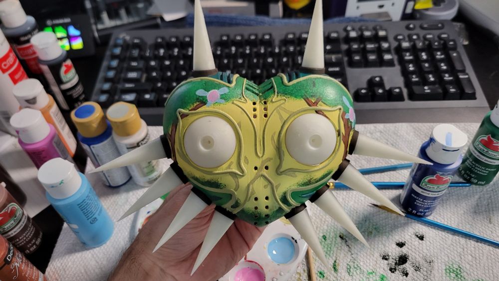 A small 3d printed majoras mask painted in yellows and greens showing a foret scene with faries