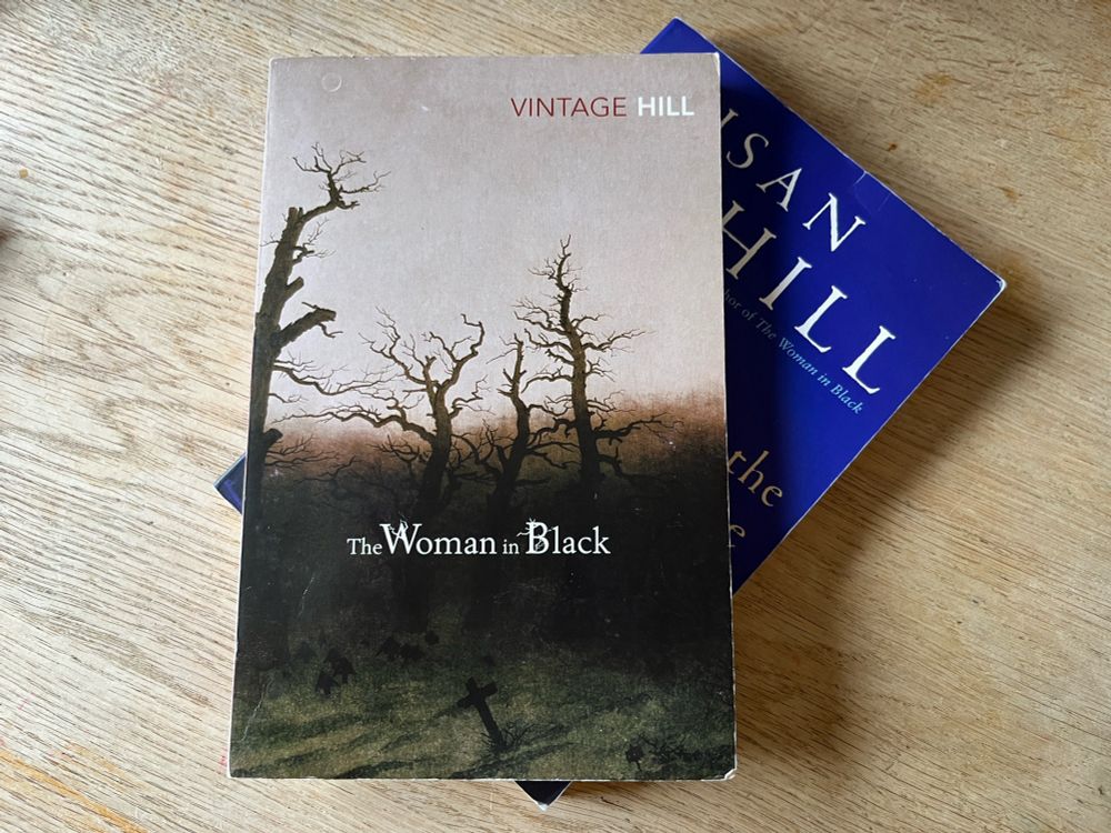 Two paperback books stacked on top of each other - barely shown is The Man in the Picture, on top is The Woman in Black, both by Susan Hill. 